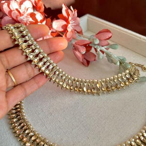 May include: Gold-toned jewelry set featuring a necklace and bracelet. The pieces are adorned with teardrop-shaped embellishments and small, round accents. The jewelry is displayed on a light-colored surface, with pink and red flowers in the background.
