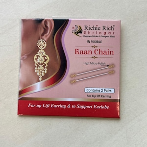 May include: A pink box with the text "Richie Rich Shringar" and "Raan Chain." The box features an image of a gold earring and contains two pairs of earring supports. The text "For up Lift Earring & to Support Earlobe" is also visible.