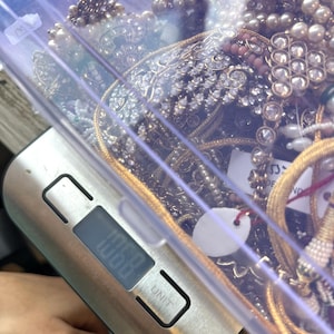 May include: A clear plastic container filled with various pieces of jewelry, including necklaces, bracelets, and earrings. The jewelry features pearls, rhinestones, and gold-colored metal. A digital scale displays a weight of 106.8 units.