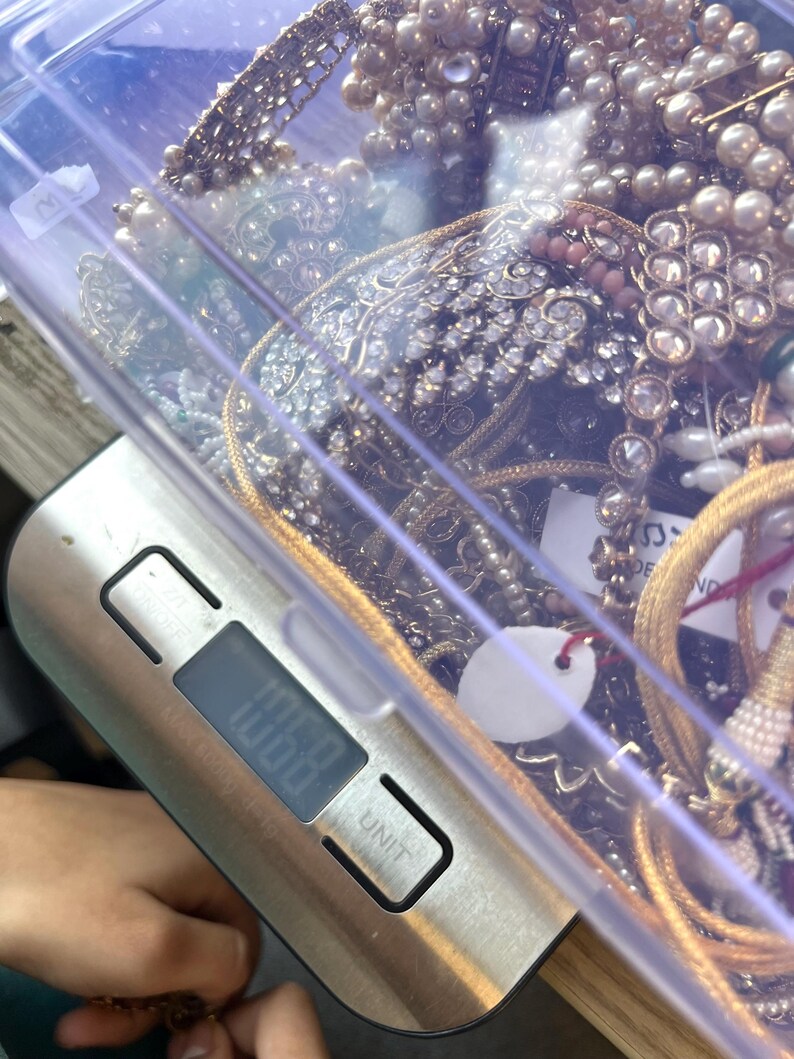 May include: A clear plastic container filled with various pieces of jewelry, including necklaces, bracelets, and earrings. The jewelry features pearls, rhinestones, and gold-colored metal. A digital scale displays a weight of 106.8 units.