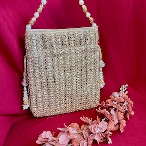 May include: A gold-coloured, woven handbag with a beaded handle and tassel accents. The bag is set against a red background, with a decorative floral arrangement in shades of pink and peach.