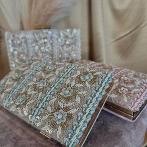 May include: Three beaded clutches in different colours. The top clutch is white with iridescent sequins. The middle clutch is beige with gold, green, and white sequins. The bottom clutch is pink with white sequins.