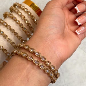 May include: A set of gold bangles with clear teardrop-shaped stones. The bangles are stacked on a person's wrist.