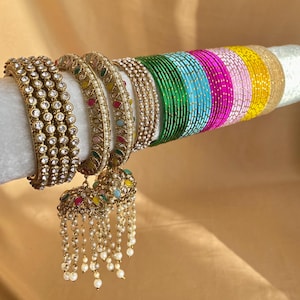 Op de afbeelding: Een collectie armbanden bestaande uit drie brede goudkleurige armbanden met heldere stenen, twee versierde goudkleurige armbanden met veelkleurige accenten en een reeks dunne, effen metalen armbanden in groen, blauw, roze, wit en geel.