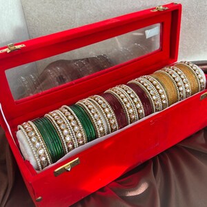 Pode incluir: Um conjunto de pulseiras empilhadas em tons de verde, bordô e dourado, cada uma com uma fileira de pedras brancas e pequenas pérolas. As pulseiras estão dispostas num suporte cilíndrico dentro de uma caixa retangular de veludo vermelho.
