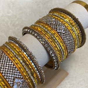 May include: A set of yellow bangles featuring metallic gold-tone bands, intricate clear rhinestone pavé detailing, and square-shaped crystal accents. The bracelets are arranged in a stack, showcasing a combination of smooth and textured surfaces.