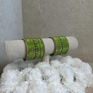 Op de afbeelding: Twee sets limoengroene armbanden met dunne, goudkleurige metalen banden afgewisseld met rijen kleine, heldere, reflecterende kristallen. De armbanden zijn in een stapel gerangschikt en vertonen een levendige, uniforme kleur en textuur.