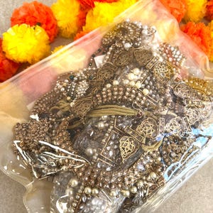 May include: A clear plastic bag filled with antique gold-toned jewellery, including necklaces, bracelets, and earrings. The jewellery is embellished with pearls and clear gemstones. Bright orange and yellow marigold flowers are in the background.