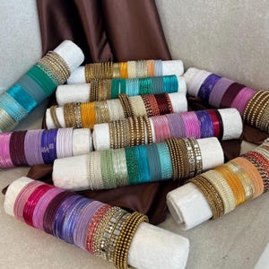 May include: A collection of numerous metal bangles arranged on white cylindrical holders. The bangles feature a variety of colors including pink, purple, green, blue, gold, and orange, with some displaying textured, beaded, or metallic finishes.
