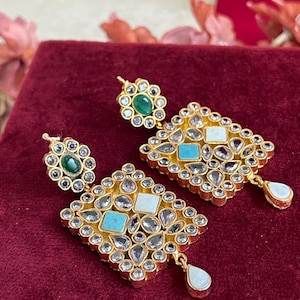 May include: A pair of gold-toned earrings with a floral stud and a diamond-shaped drop. The earrings feature turquoise and white square stones, surrounded by clear, round stones. A pear-shaped stone hangs from the bottom of each earring.