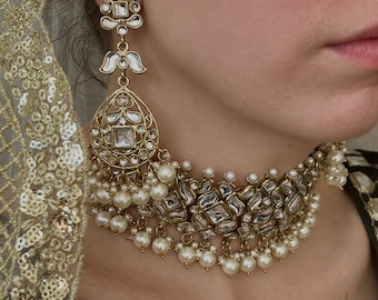 May include: Close-up of a gold-toned necklace and earring set. The jewelry features clear, square-cut stones, white enamel accents, and dangling faux pearls. The necklace is a choker style.