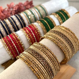 May include: A collection of stacked bangles arranged on cylindrical holders. The sets feature various colours including gold, red, dark blue, and green, with intricate metallic textures, beaded details, and sparkling accents on each individual bracelet.