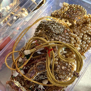 May include: A clear plastic container overflowing with gold-toned jewelry. The jewelry includes necklaces, bracelets, and earrings, featuring pearls, rhinestones, and intricate designs. A gold rope-like necklace is in the foreground.
