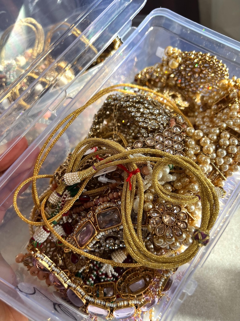 May include: A clear plastic container overflowing with gold-toned jewelry. The jewelry includes necklaces, bracelets, and earrings, featuring pearls, rhinestones, and intricate designs. A gold rope-like necklace is in the foreground.