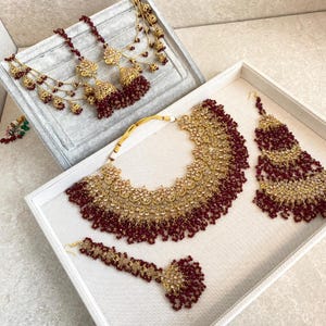 Peut inclure: Un ensemble de bijoux de mariée en or et bordeaux avec un collier imposant, des boucles d'oreilles assorties et un tikka. Le collier présente des motifs floraux complexes et est orné de pierres scintillantes.
