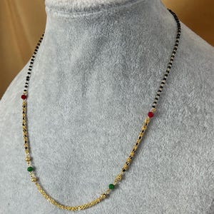 May include: A gold chain necklace with black beads and red and green accents. The necklace is on a grey background.