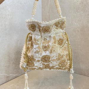 May include: Ivory-colored drawstring bag with gold floral embroidery and sequin accents. The bag features pearl beaded handles and tassel details. A luxurious, ornate accessory.