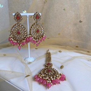 May include: A set of gold-tone earrings and a matching tikka, featuring pink and white beads and clear stones. The jewelry is displayed on a white stand against a white and gold background.