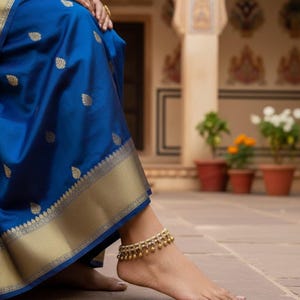 May include: A close-up of a woman's foot adorned with a gold anklet featuring dangling charms. The foot rests on a stone surface, with a vibrant blue fabric with gold accents in the background. The scene evokes a sense of elegance and cultural richness.