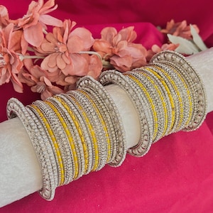 May include: A set of stacked bangles featuring alternating bands of bright yellow and metallic silver. The silver bangles are adorned with small, white, bead-like textures, while the yellow bands have a smooth, solid finish. The bangles are arranged in a repeating pattern on a cylindrical display.
