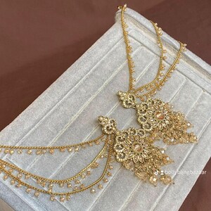 May include: Gold-toned, ornate earrings with floral and dangling bead details. The earrings are displayed on a light gray surface. The design includes multiple chains and small, sparkling beads, creating a luxurious look.
