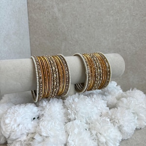 May include: Two sets of gold-toned bangles featuring a combination of smooth metallic bands and textured bands adorned with small white faux pearls and clear crystals. The bangles are arranged in a stacked formation on a cylindrical display stand.