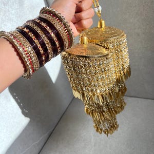 May include: A set of dark maroon and gold bangles featuring intricate crystal embellishments, paired with a tiered gold kalira ornament. The kalira consists of multiple layers of metallic gold beads, small dangling leaf-shaped charms, and a dome top.
