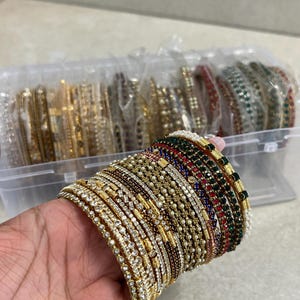 May include: A collection of stacked bangles featuring gold-tone metal, clear rhinestones, small white faux pearls, and alternating red, green, and blue faceted stones. The set includes various textured designs and is stored in a clear plastic box.