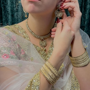 May include: A woman wearing a white and gold embroidered saree with a gold necklace and earrings. She is also wearing gold bangles on her wrists.