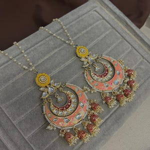 May include: A pair of elaborate earrings with a coral and red crescent moon design, adorned with clear stones and dangling red and gold bell charms. The earrings have a yellow floral top and are displayed on a grey surface.