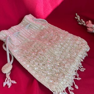 May include: An ivory and silver beaded drawstring bag. The bag is covered in small, clear beads, with a fringe of beads along the bottom edge. The drawstring is a silver cord with a decorative tassel. The bag is set against a red background.