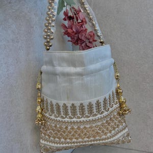 May include: A white handbag with gold embroidery and beaded handles. The bag has a clear plastic top and is filled with pink flowers. Gold tassels hang from the sides of the bag.