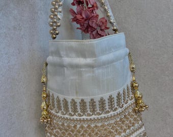 Könnte beinhalten: Eine weiße Handtasche mit goldener Stickerei und Perlenhenkeln. Die Tasche hat einen transparenten Kunststoffaufsatz und ist mit rosa Blumen gefüllt. Goldene Quasten hängen an den Seiten der Tasche.