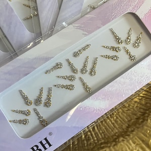 Puede incluir: Una colección de pedrería para uñas en una bandeja blanca. La pedrería está dispuesta en un diseño floral con bases doradas y cristales transparentes y brillantes. La bandeja está dentro de una caja morada y blanca.