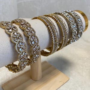 May include: A collection of gold-coloured bangles, adorned with clear, sparkling stones, are presented on a white, curved display stand. The bangles showcase diverse designs, including floral and linear patterns, offering a variety of textures and styles. The stand rests on a wooden base.