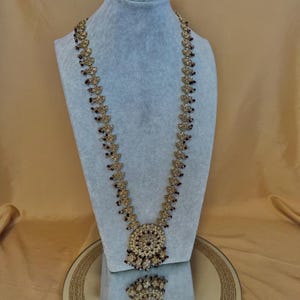 May include: A gold-coloured necklace and pendant set displayed on a blue mannequin. The necklace has a long chain with leaf-shaped details and small, dark red beads. The pendant is a large, circular design with similar red beads.