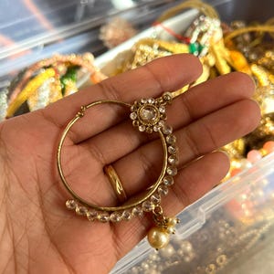 May include: A gold-toned nose ring with a circular design. The ring features a central clear gemstone surrounded by smaller clear stones and a dangling pearl. The jewelry is held in a hand, with other jewelry visible in the background.