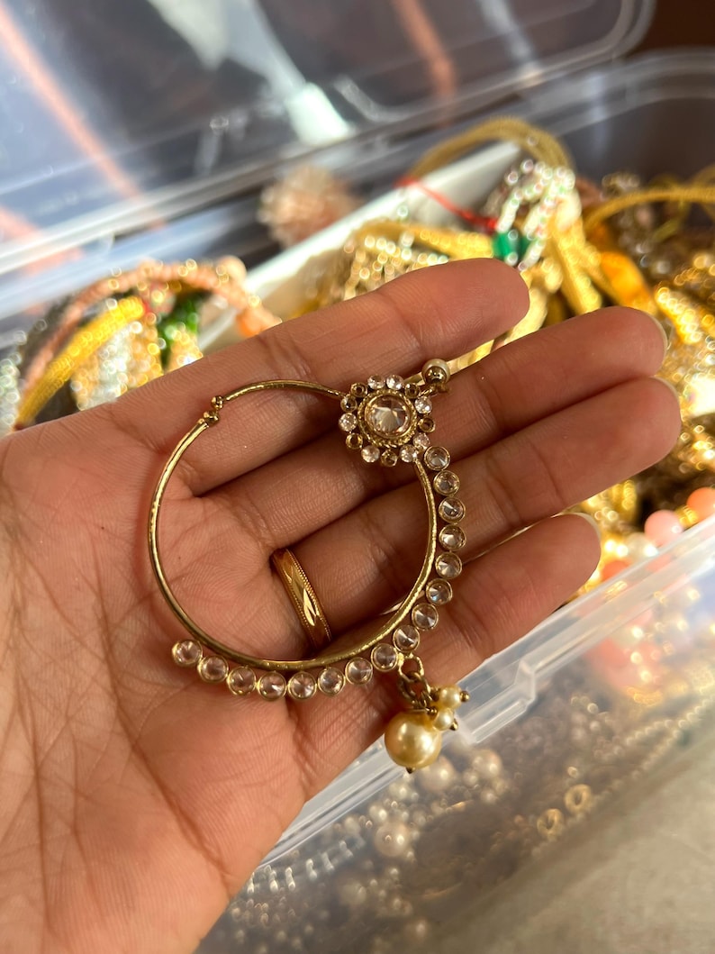 May include: A gold-toned nose ring with a circular design. The ring features a central clear gemstone surrounded by smaller clear stones and a dangling pearl. The jewelry is held in a hand, with other jewelry visible in the background.