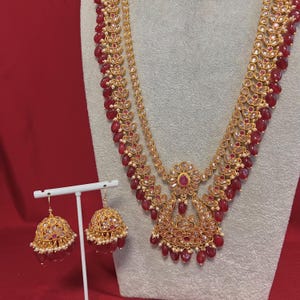 Könnte beinhalten: Goldfarbenes Halsketten- und Ohrring-Set mit roten Edelstein-Akzenten. Die Halskette hat mehrere Stränge und einen Anhänger mit floralem Design. Die Ohrringe sind glockenförmig mit passenden roten Perlen. Das Schmuckstück wird auf einem weißen Ständer präsentiert.