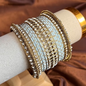 May include: A set of stacked bangles featuring a combination of light blue and gold-toned metal bands. The design includes intricate gold filigree patterns and rows of small, clear, sparkling rhinestones embedded along the edges and centre.