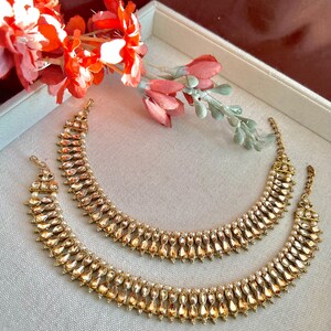 May include: Two gold-toned anklets with a teardrop and pearl design. The anklets are displayed on a light gray surface, with red and pink flowers in the background. The jewelry has a vintage or antique aesthetic.