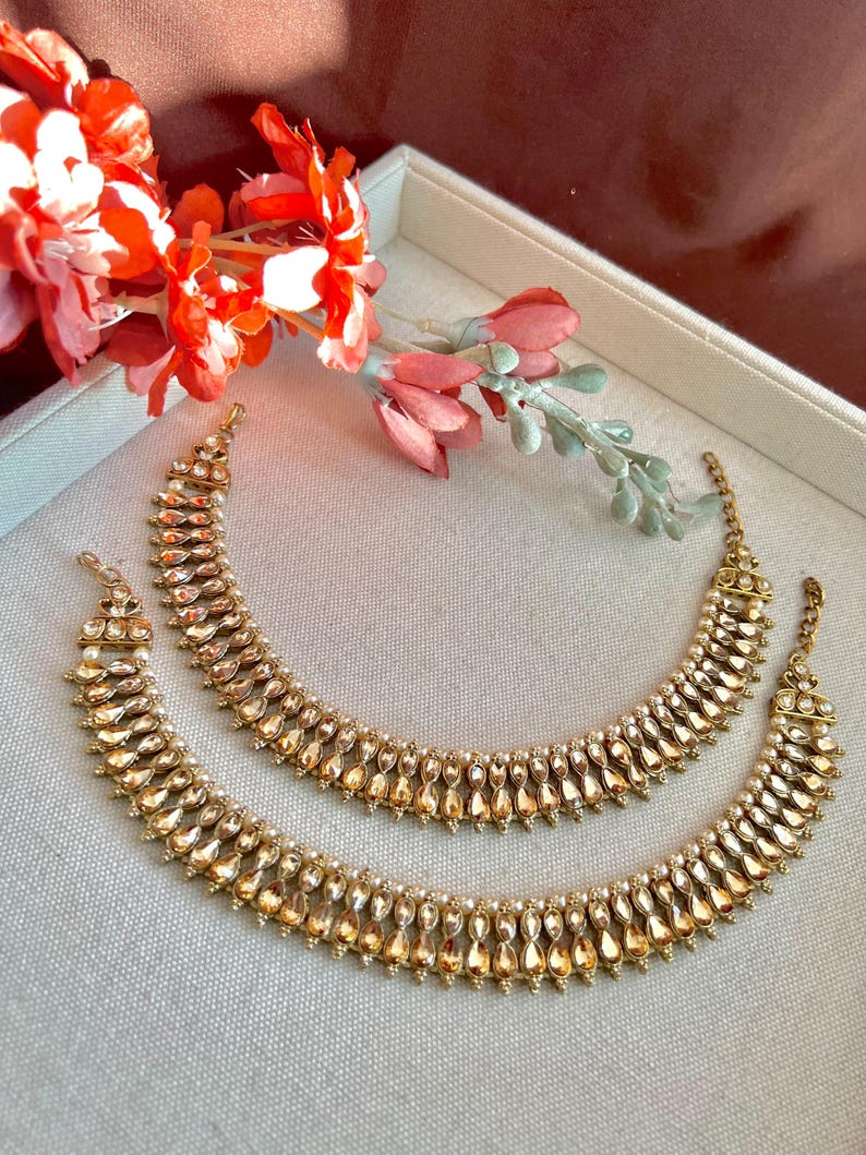 May include: Two gold-toned anklets with a teardrop and pearl design. The anklets are displayed on a light gray surface, with red and pink flowers in the background. The jewelry has a vintage or antique aesthetic.