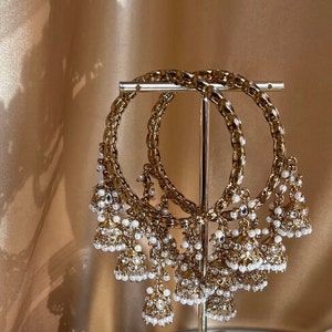 May include: Gold bangles with detailed chain links and hanging bell charms. Each charm features small, clear crystals and clusters of white pearls, creating an elegant design. The jewellery is displayed on a gold stand, showcasing the ornate details.