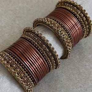 Peut inclure: Un ensemble de bracelets joncs composé de fines bagues métalliques couleur cuivre alternées avec des bandes dorées texturées, incrustées de petits cristaux marron clair et de minuscules perles blanches, disposés en pile cylindrique.