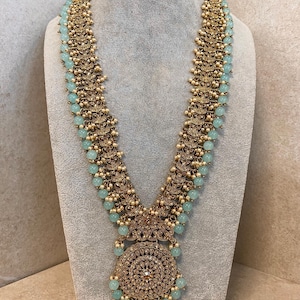 Puede incluir: Un collar dorado con un gran colgante y cuentas azul claro. El collar se exhibe en un maniquí gris. El colgante tiene un diseño circular con detalles intrincados y una gema central.