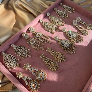 May include: A collection of gold tone dangle earrings with clear and amber coloured crystals. The earrings are displayed on a pink velvet surface.