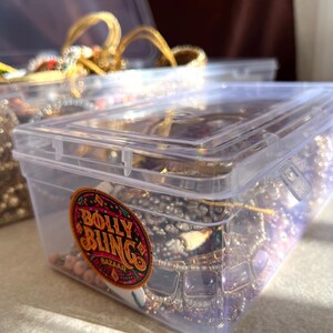 May include: Clear plastic storage containers filled with jewelry. The containers have lids and are filled with necklaces, bracelets, and other accessories. One container has a sticker that says "Bolly Bling Bazaar".