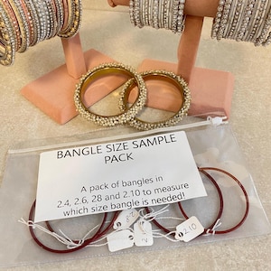 May include: A selection of bangles in different designs and sizes, presented on pink display stands. A clear plastic bag is visible, marked "BANGLE SIZE SAMPLE PACK", containing red bangles and size tags. The text on the bag reads: "A pack of bangles in 2.4, 2.6, 28 and 2.10 to measure which size bangle is needed!"
