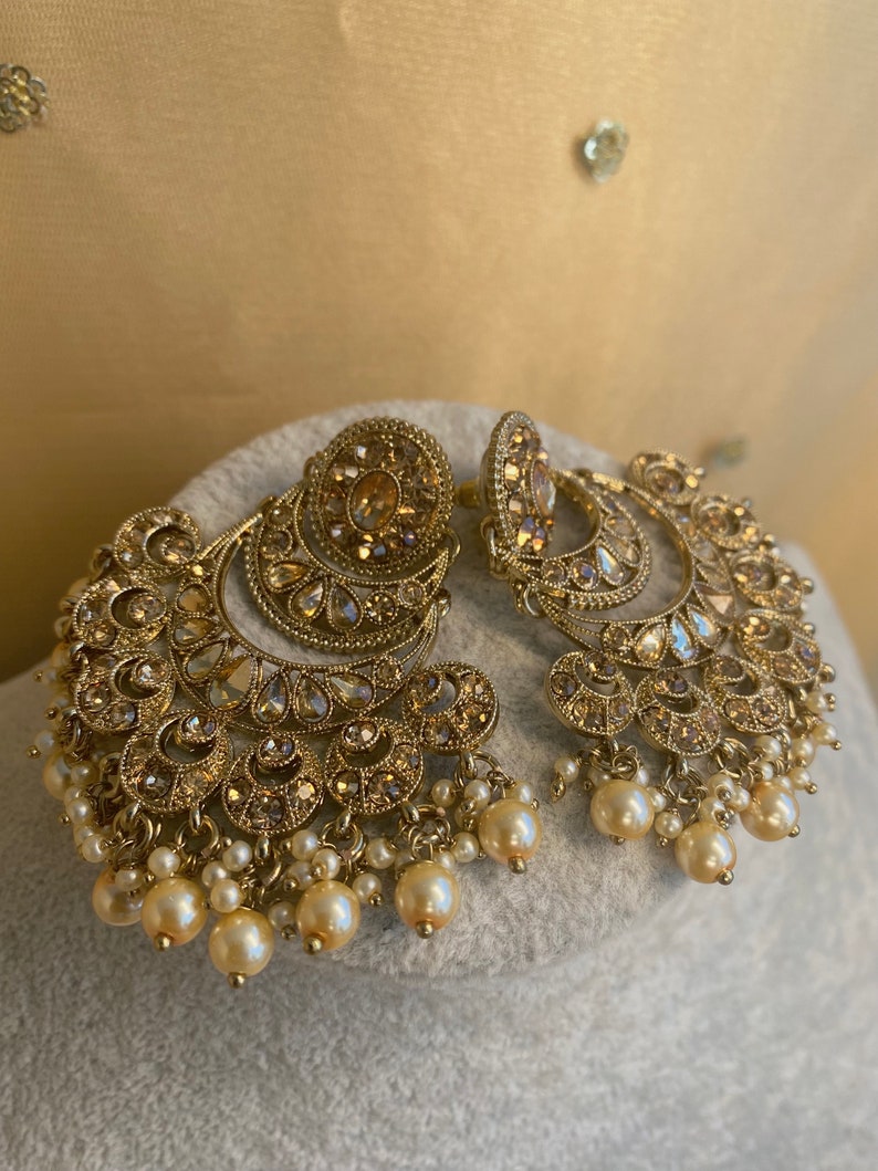 Peut inclure: Une paire de boucles d'oreilles en or avec un motif de croissant de lune et des accents de perles. Les boucles d'oreilles sont orn&eacute;es de strass scintillants.