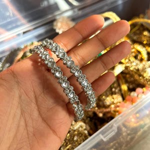 May include: Two silver bangles with heart-shaped clear stones are held in a hand. The bangles are in focus, with other jewelry visible in the background. The jewelry is stored in a clear plastic container.